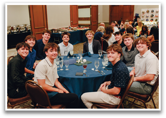 A group of young men are sitting around a table with wine glasses, forks, knives, and spoons. They are posing for a picture. Contenido generado con IA