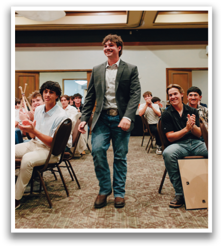 A man in a suit and tie stands in a room with other people. He is holding a drum and clapping. Contenido generado con IA