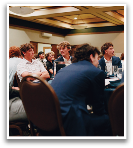 A group of people are sitting in a room, some wearing suits. They are seated around a table with wine glasses and cups. There are several chairs in the room, some occupied and others empty. The people are engaged in conversation and enjoying their time together. Contenido generado con IA