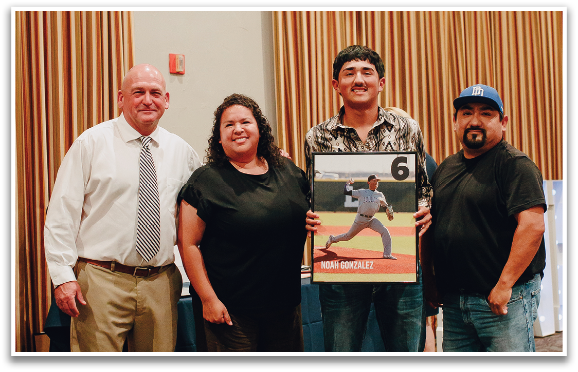 Three people are standing in front of a stage, posing for a picture. One of the men is holding a baseball bat. Contenido generado con IA