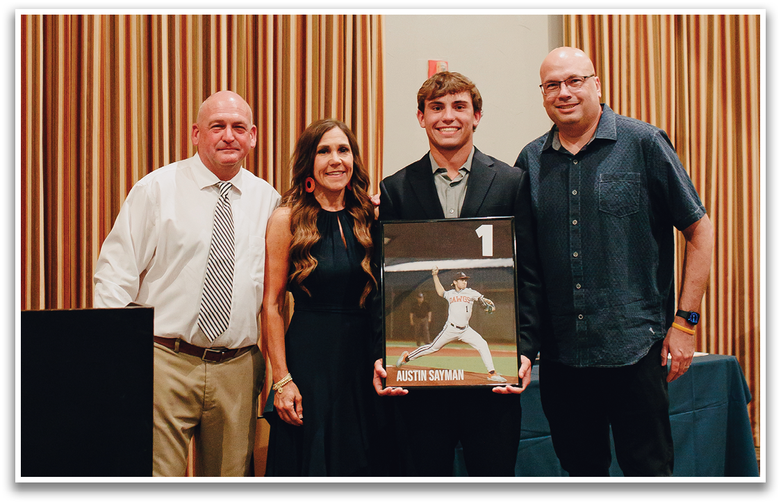 Three people are standing in front of a picture, with a man holding a framed item. The man on the left is wearing a tie, and the woman on the right is wearing a dress. The man in the middle is holding the framed item, which appears to be a baseball. Contenido generado con IA