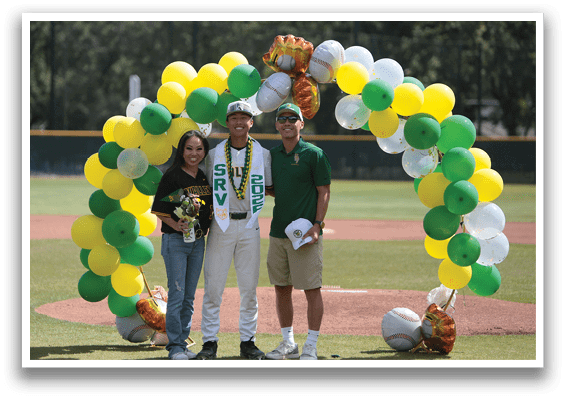 A man and woman stand in front of a baseball field with a green and yellow arch behind them. Description generated by AI
