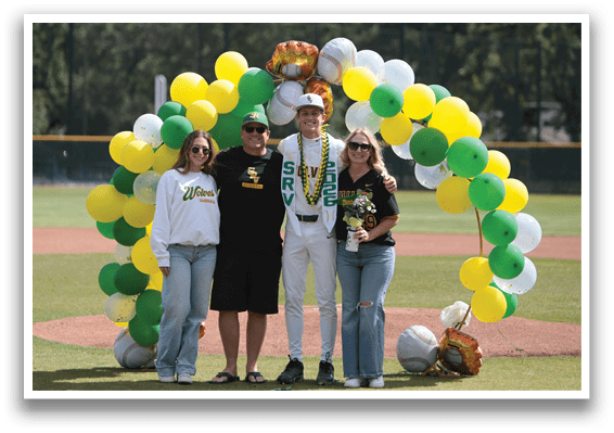 A group of people standing in front of a baseball field with a green and yellow banner. Description generated by AI