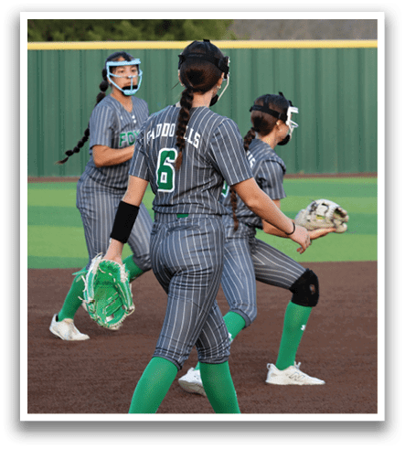Three female softball players wearing green and gray uniforms are on the field. One player is holding a catcher's mitt, another is wearing a helmet, and the third is standing in the outfield. They are all ready to catch the ball. Contenido generado con IA