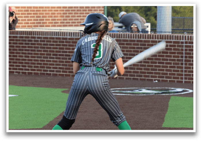 A female baseball player in a grey and black uniform is holding a bat and getting ready to swing. Contenido generado con IA