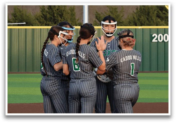 Four female softball players wearing gray uniforms and green socks are huddled together on the field. Contenido generado con IA