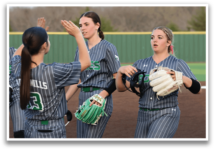 Four female softball players wearing grey and green uniforms are standing on a field. They are holding their gloves and are in the process of high-fiving each other. Contenido generado con IA