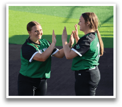 Two women are on a baseball field, one of them is wearing a green shirt and the other is wearing a black shirt. They are giving each other high fives. Contenido generado con IA