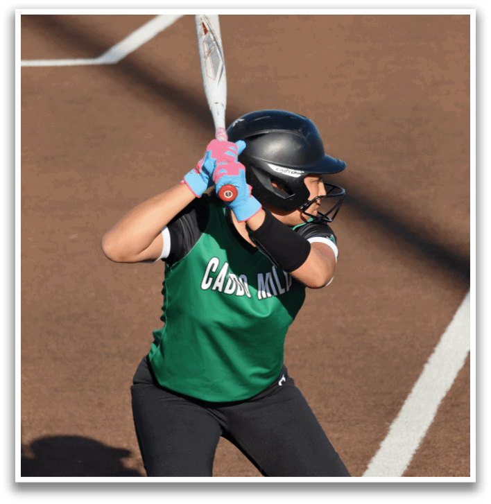 A female baseball player in a green and black uniform is swinging a bat at a ball. Contenido generado con IA