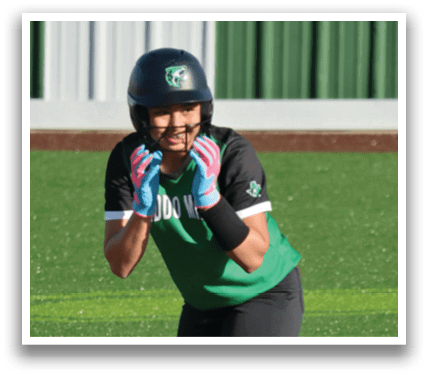 A female baseball player in a green and black uniform is standing on a field, holding her glove up. Contenido generado con IA
