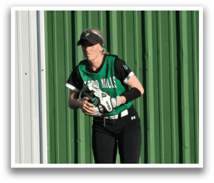 A woman wearing a green and white shirt and black pants is standing on a grassy field. She is holding a baseball in her hand. Contenido generado con IA
