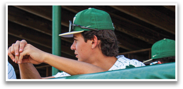 A baseball player wearing a green and white uniform stands behind a fence, pointing at something. AI generated content