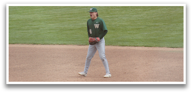 A baseball player wearing a green shirt and grey pants standing on the field. Description generated by AI