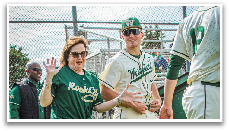 A woman wearing a green shirt and black skirt is waving at a man in a green and white baseball uniform. Description generated by AI