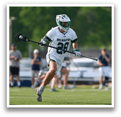 A man wearing a white and green shirt and white shorts is running with a lacrosse stick. Description generated by AI