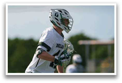 A man in a white shirt and green shorts is holding a lacrosse stick. Description generated by AI