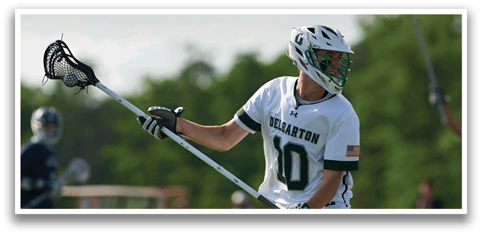 A man wearing a white shirt and green and white shorts holding a lacrosse stick. Description generated by AI