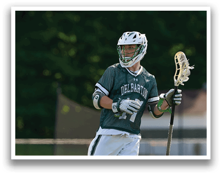 A man wearing a green and white shirt and white pants holding a lacrosse stick. Description generated by AI