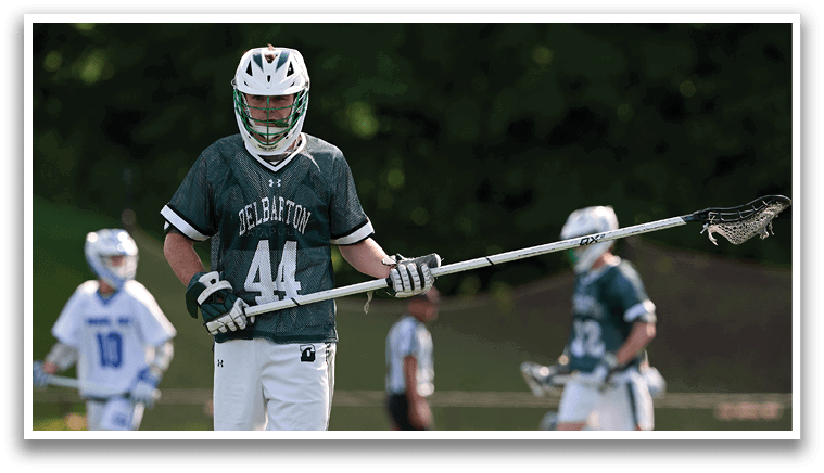 A man wearing a green shirt and white shorts holding a lacrosse stick. Description generated by AI