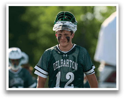 A man in a green shirt and white shorts is holding a lacrosse stick. Description generated by AI