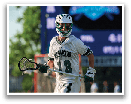 A man wearing a green and white shirt and white shorts is running with a lacrosse stick. Description generated by AI