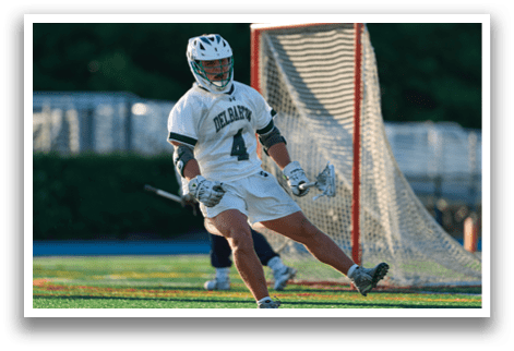 A man in a green shirt and white shorts is holding a lacrosse stick. Description generated by AI