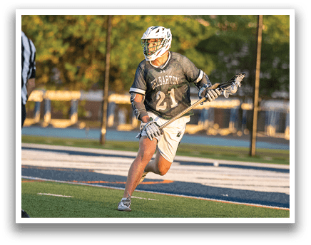 A man in a green shirt and white shorts is holding a lacrosse stick. Description generated by AI