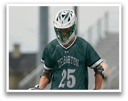 A man wearing a green and white shirt and white pants holding a lacrosse stick. Description generated by AI