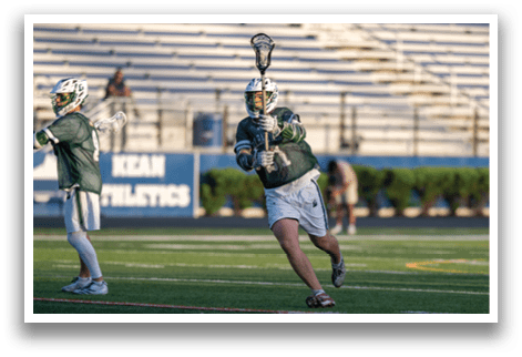 A man in a green shirt and white shorts is holding a lacrosse stick. Description generated by AI