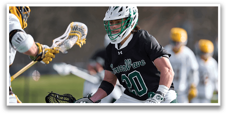 A man wearing a black and green shirt and white shorts holding a lacrosse stick. Description generated by AI