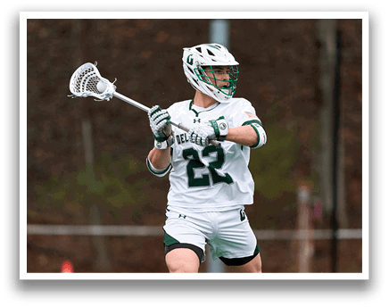 A man wearing a white shirt and green shorts is holding a lacrosse stick. Description generated by AI