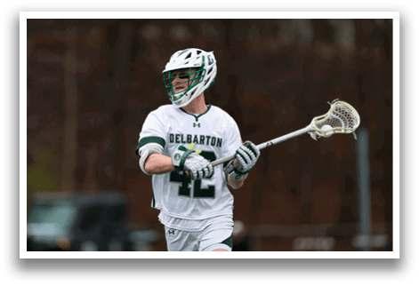 A man wearing a white shirt and green shorts holding a lacrosse stick. Description generated by AI