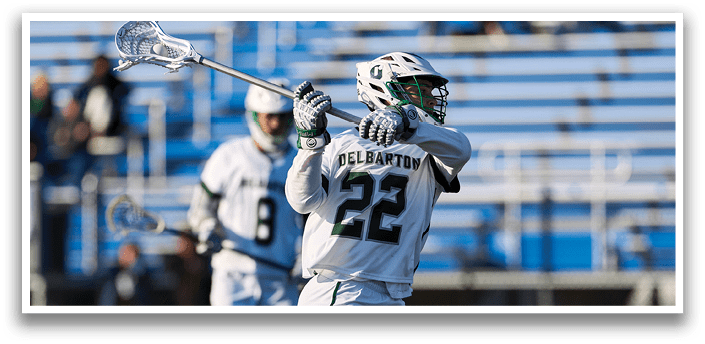 A man wearing a white and green jersey is holding a lacrosse stick. Description generated by AI