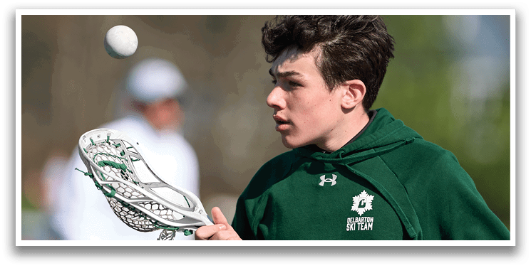 A man in a green hoodie is holding a lacrosse stick and looking up at a ball. Description generated by AI
