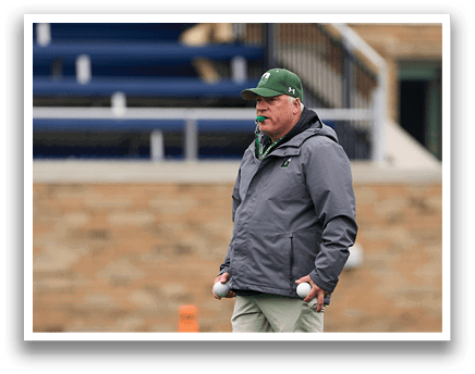 A man wearing a gray jacket and green hat standing on a field. Description generated by AI