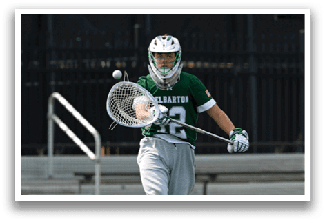 A man wearing a green and white shirt and grey pants is playing lacrosse. Description generated by AI