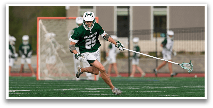 A man wearing a green and white jersey is running on a field with a lacrosse stick. Description generated by AI