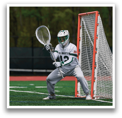 A man wearing a white and green shirt and grey pants is playing lacrosse. Description generated by AI