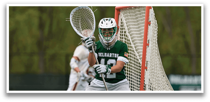 A man wearing a green and white shirt and grey pants is holding a lacrosse stick. Description generated by AI