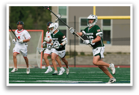 A man with a green shirt and white shorts is holding a lacrosse stick. Description generated by AI