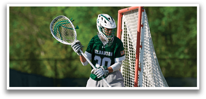 A man wearing a green and white shirt and grey pants is holding a lacrosse stick. Description generated by AI