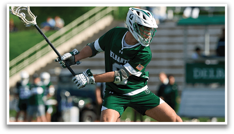 A man wearing a green and white uniform is holding a lacrosse stick. Description generated by AI