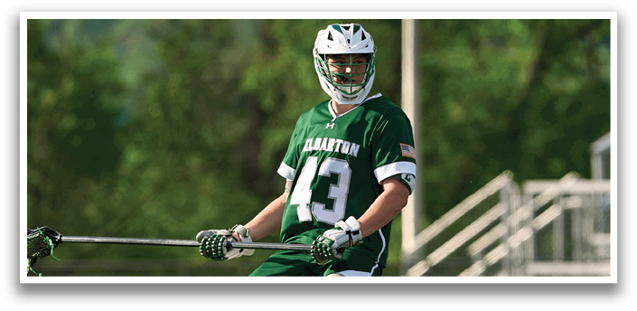 A man wearing a green and white jersey is holding a lacrosse stick. Description generated by AI