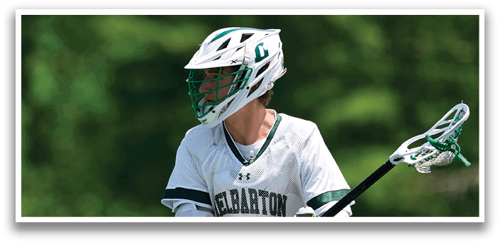 A man wearing a white shirt and green and white helmet holding a lacrosse stick. Description generated by AI