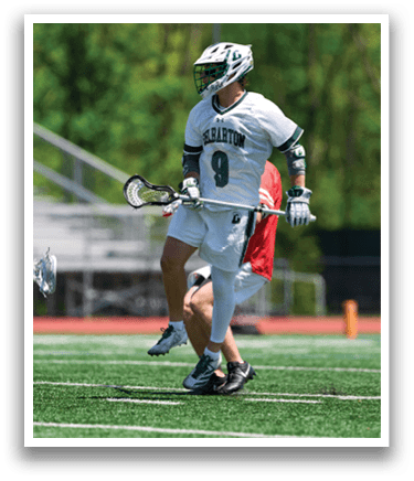 A man wearing a white shirt and green and white shorts is playing lacrosse. Description generated by AI