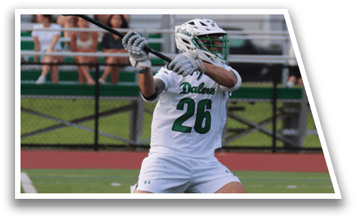 A man wearing a white uniform and green number 26 is playing lacrosse. Description generated by AI