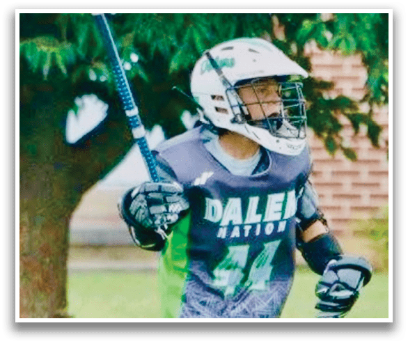 A young man wearing a grey and green jersey is holding a lacrosse stick and walking on a grassy field. AI generated content