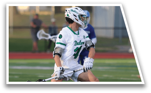 A man wearing a white shirt and green shorts is holding a lacrosse stick. Description generated by AI