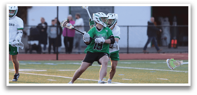 A man in a green shirt and black shorts is holding a lacrosse stick. Description generated by AI
