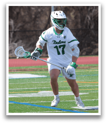 A man in a white shirt and green shorts is holding a lacrosse stick and walking on a field. Description generated by AI